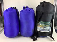 Three sleeping bags packed in stuff sacks: two purple and one black with a label.