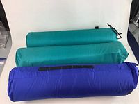 Three rolled camping inflatable sleeping mats in blue and green storage bags lying on white surface.
