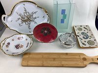 Seven assorted items including fine china dishes, glass vase, condiment jar, and bamboo board arranged on white background