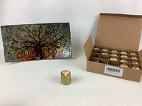 Set of 16 gold glitter LED candles in open cardboard box beside one outside candle and the decorative tree plate on white table.