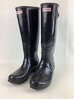 Front and side view of pair of black Hunter tall rain boots with brand logo at top cuff, showing overall condition and scuffs.