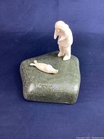 Full view of sculpture showing green polished stone base with two small white figures, one standing human figure with hooded clothing and one seal lying on the base.