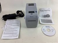Frontal view of Intermec PC23d printer with power adapter, manuals and software CD laid out on table.