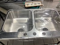 Top-down view of new stainless steel double sink wrapped in protective plastic, showing two deep basins and faucet holes