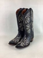 Pair of women's black leather cowboy boots with intricate white embroidery and studs shown standing side by side from a front-side angle.
