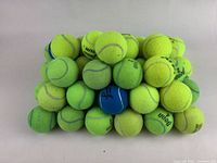 Pile of about 50 used tennis balls, mostly yellow with a few blue ones, arranged in a pyramid shape on white background.