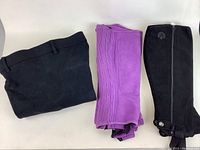 Three equestrian chaps and half chaps displayed on a white surface: one full pair of black riding chaps folded, one purple pair of half chaps, and one black pair of half chaps.