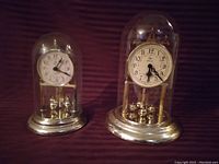 Two anniversary clocks, each with white clock faces, Arabic numerals, gold-tone bases, and four ball pendulums, enclosed in glass domes