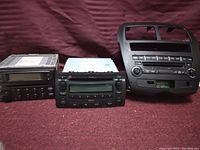 Three automotive stereo head units with AM/FM radio and CD players shown side by side on a burgundy fabric surface