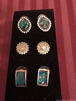 Photo showing three pairs of stud earrings on a black velvet display, with varying shapes and colors including green iridescent, floral with pink and yellow stones, and rectangular green-blue stones.
