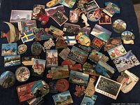 Top view of over 60 assorted souvenir magnets from various countries placed on a dark blue fabric background. Includes magnets with scenes, flags, animals, and building designs.
