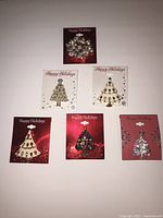 Six distinct holiday-themed Christmas tree pins on display cards with Happy Holidays text, laid out on a white background.