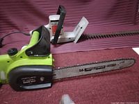 Photo showing a Portland 14-inch electric chainsaw with green and black casing next to a white and black electric hedger, placed on a red surface with a striped background.
