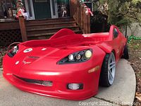 Left angled view showing detailed front end and side wheel design of the red Corvette bed frame outdoors on pavement.