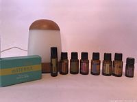 Photo of diffuser, 7 essential oil bottles in amber glass with black caps, and DoTerra soap bar in teal/yellow box on pink background