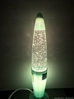 Glitter lava lamp illuminated against a wall showing green glow and floating silver glitter in clear liquid.