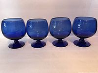 Four deep cobalt blue hand blown glass goblets displayed in a row, showing rounded bowls, short stems, and flat wide bases with pontil marks.