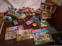 Photo showing a large collection of LEGO Friends sets including a cruise ship, several cars, a camper, a three-story building with balconies and a pool scene among other structures, along with LEGO Friends minifigures scattered throughout. Several instruction booklets for different sets are placed in front of the models.
