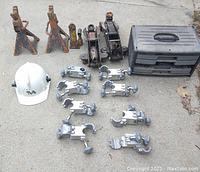 Photo showing two orange jack stands, two hydraulic jacks (black and silver), two bottle jacks, eight silver clamps, white hard hat, and black plastic tool case on concrete floor.