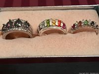 Photo of three silver tone rings with various multicolored gemstones displayed on beige velvet holder.