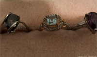 Close-up front view of three rings with different colored gemstones on a light tan cushioned holder.