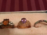 Photo of three rings on cushioned surface: silver ring with orange gemstone and clear stones, gold-tone ring with purple gemstone, thin band with multicolored stones