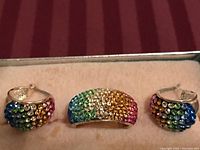 Photo showing a set of three jewelry items consisting of a multicolored rhinestone ring and two matching hoop earrings inside a jewelry box with a tan interior and a red striped fabric background.