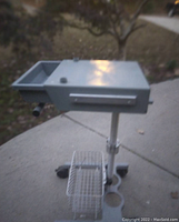 Side view of the gray metal medical cart showing the locking top lid, drawer below, and wire basket under the drawer, mounted on a heavy wheeled base.