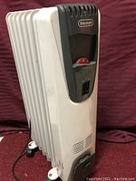 White and gray DeLonghi oil-filled electric space heater showing front controls and radiator fins, placed on a red fabric surface.