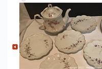 Photo showing a white porcelain teapot with lid and five matching dessert plates. Plates have scalloped rims with gold trim and floral designs. Teapot matches with similar floral pattern.