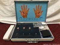Device displayed inside open metal case with hand acupoint diagrams on lid. Three knobs and four output jacks visible alongside wires and electrode pads.