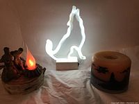 Photo of all three items: neon wolf light, campfire light with figurines, and scenic candle together on white cloth.