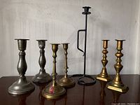 Photo showing all seven candlesticks arranged side by side on a wooden surface against a white wall, illustrating the variety of materials and sizes.