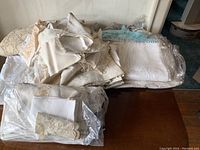 Overview photo showing a large pile of folded cream and white linen tablecloths and napkins with delicate embroidery and scalloped edges, and a wrapped set in plastic.