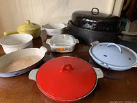Photo showing the full collection of vintage casseroles and pans including the red Voss Denmark casserole in front, blue and white dishes, yellow pot, clear glass dish with flowers, and large black roasting pan.