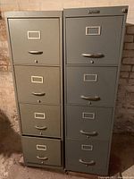 Two gray metal four-drawer file cabinets side by side against a white brick wall, showing front faces with metal handles and label holders.