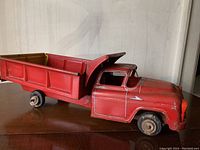 Side view of red vintage tin toy dump truck showing metal construction, wheels, and cab details.