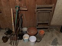 Photo showing assortment of garden tools including shovel, garden fork, pruning shears, watering can, several planters of terracotta and metal, and a wooden foldable chair against wooden wall.