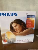 Front of box showing the Philips Wake-Up Light and a woman lying down, with features described such as colored sunrise simulation, 5 natural sounds, FM radio, fall asleep function, bedside light with 20 light settings, and tap snooze.