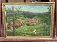 Full view of framed valley landscape oil painting showing houses, trees and two figures in foreground. Frame has visible chips.