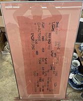 Photo of framed Asian print artwork showing the full piece with black symbols on a red background, displayed on the floor among other items.