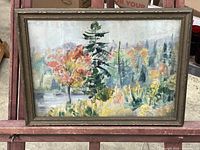 Framed oil painting showing a natural landscape with trees and colorful foliage, set on a red wooden slatted chair.