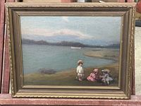 Front view of the oil painting showing three children near a lake with boat and mountains in distance, framed in decorative gold-tone frame with some visible chipping.