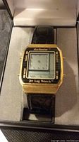 Front view of Acclimator Jet Lag Watch showing dual time LCD display, gold-tone bezel, and black leather strap inside original box.
