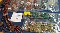Overview of 7 polished stone beaded necklaces in various colors, arranged on a blue background, some in sealed plastic bags. Several have silver-tone floral and decorative bead accents.