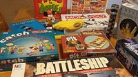 Photo showing an assortment of board games including Catch Phrase, Monopoly, Pit, Mancala, MindTrap, Battleship, plus a Mickey Mouse plush, poncho, kaleidoscope, and a crystal growing kit.