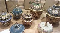 Seven Thai ceramic trinket boxes together, showing various traditional gold and colorful hand painted designs on lids and bodies