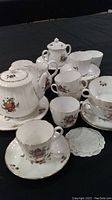 Full view of tea set pieces including teapot, creamer, sugar bowl, cups, and saucers arranged on black surface, showing overall set and pattern details.