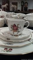 Close-up image showing cup, saucer, and plate with fruit and floral pattern, and gold rim detail on fine porcelain material.