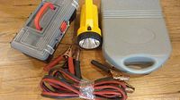 Photo of gray and red tool case, yellow and black flashlight, and red/black jumper cables on wood floor